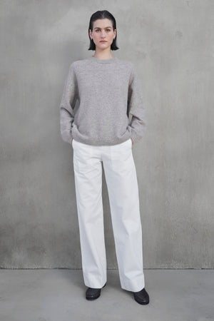 A person with short dark hair stands against a gray wall, wearing the LAKEWOOD ALPACA SWEATER by Velvet by Jenny Graham, white wide-leg pants, and black shoes. The person's hands are tucked into their pockets.