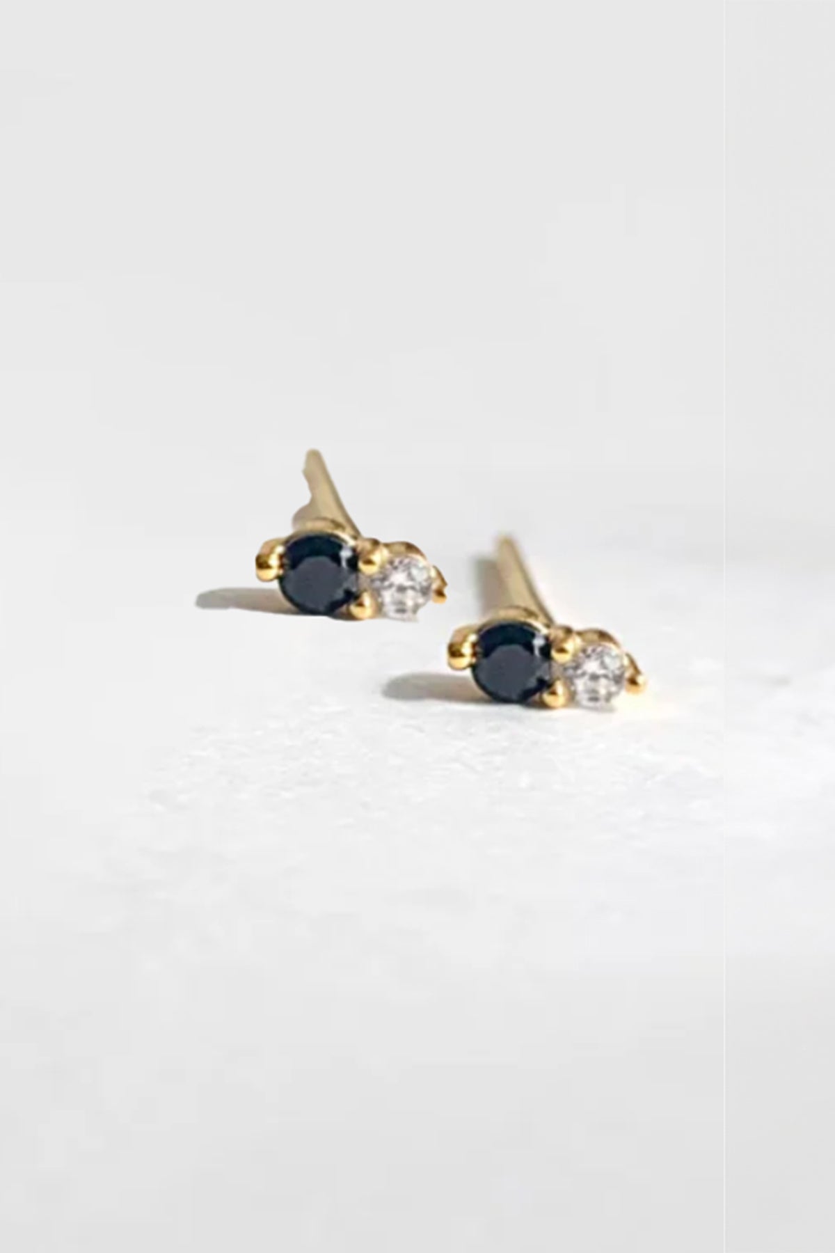 Close-up of the "Twin Stud Onyx" earrings by Phyllis+Rosie, showcasing small stud earrings with gold settings featuring one black and one clear gemstone on each. These handcrafted pieces evoke the allure of vintage jewelry, drawing inspiration from New York City's pop and vintage street culture, presented against a plain grey background. *Excluded from discounts/promotions.-37930792812737
