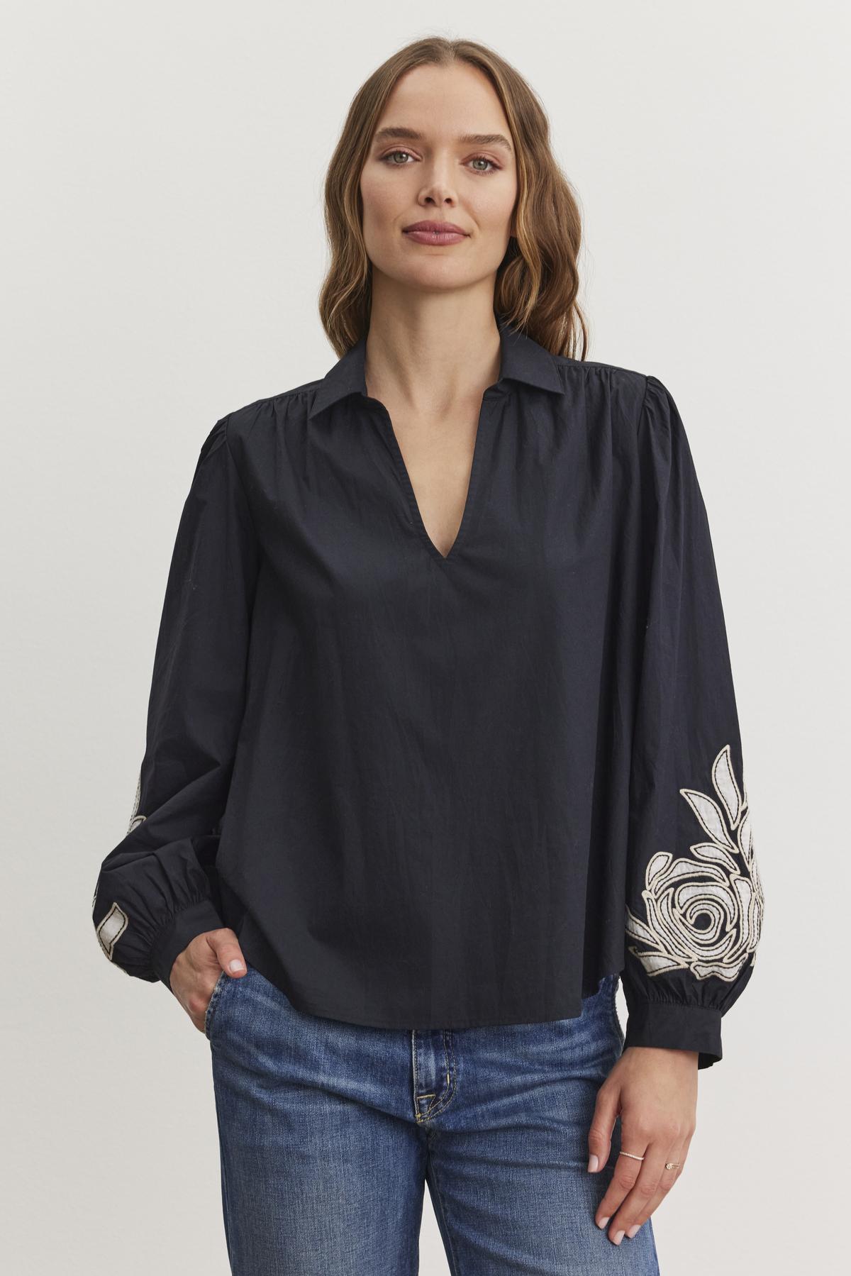 A person with long hair wears the Velvet by Graham & Spencer MARTINE TOP, a dark blouse with a V-neckline and two-tone embroidered sleeves, paired with blue jeans. They stand against a plain background.-38512796827841