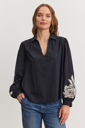 A person with long hair wears the Velvet by Graham & Spencer MARTINE TOP, a dark blouse with a V-neckline and two-tone embroidered sleeves, paired with blue jeans. They stand against a plain background.