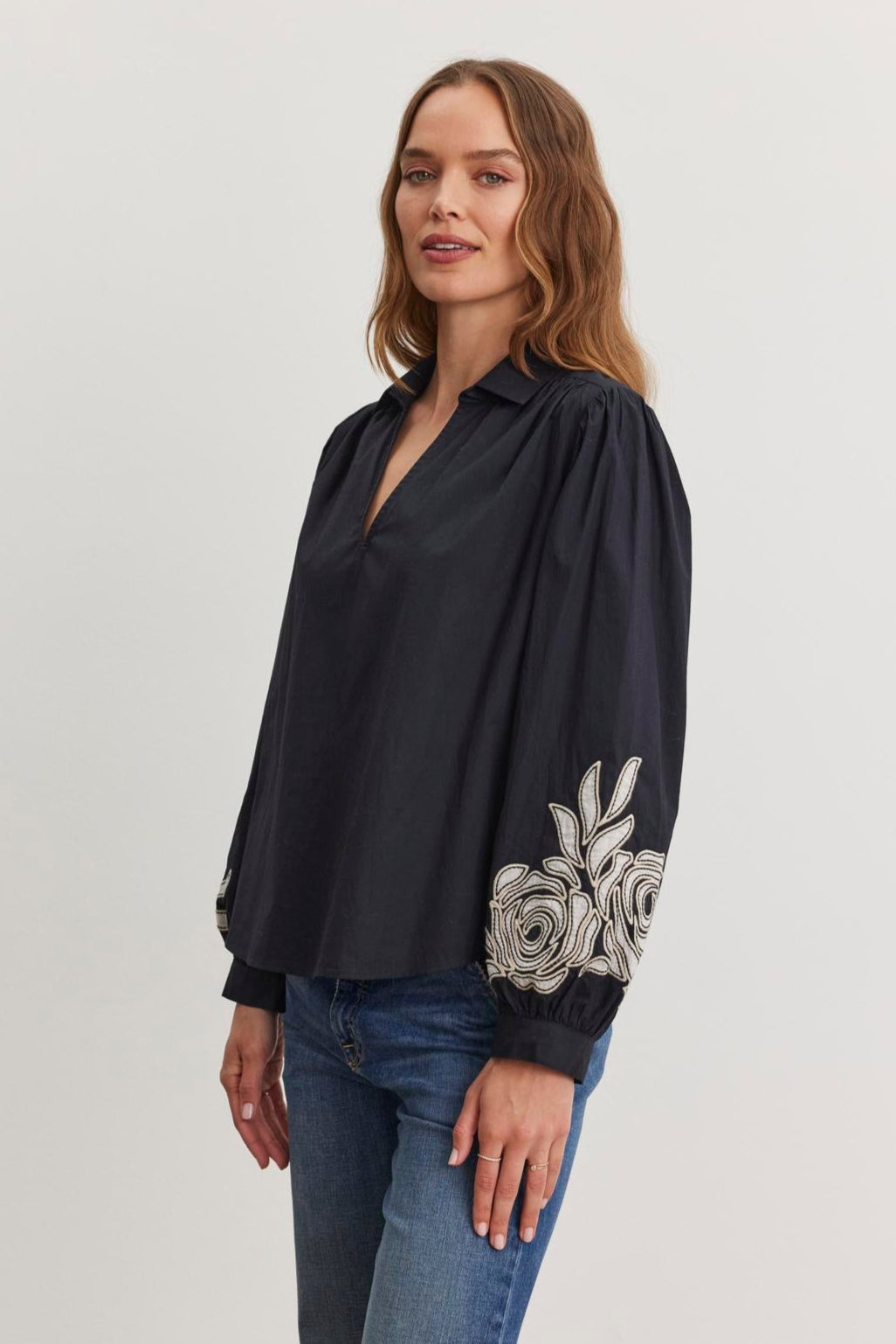 A woman wearing the Velvet by Graham & Spencer MARTINE TOP, featuring a black cotton poplin fabric with a V-neckline and two-tone floral embroidery on the sleeves, stands against a plain background, pairing it with blue jeans.-38512796893377