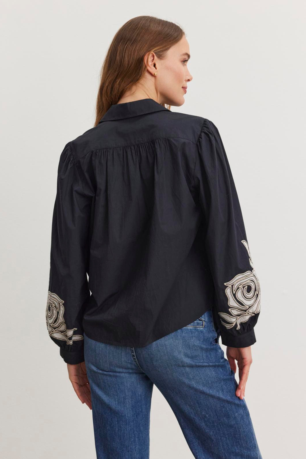 A woman wearing the Velvet by Graham & Spencer MARTINE TOP, a black cotton poplin shirt with two-tone embroidered sleeves, paired with blue jeans stands sideways against a white background.
