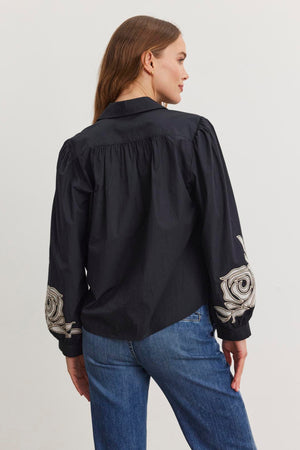 A woman wearing the Velvet by Graham & Spencer MARTINE TOP, a black cotton poplin shirt with two-tone embroidered sleeves, paired with blue jeans stands sideways against a white background.