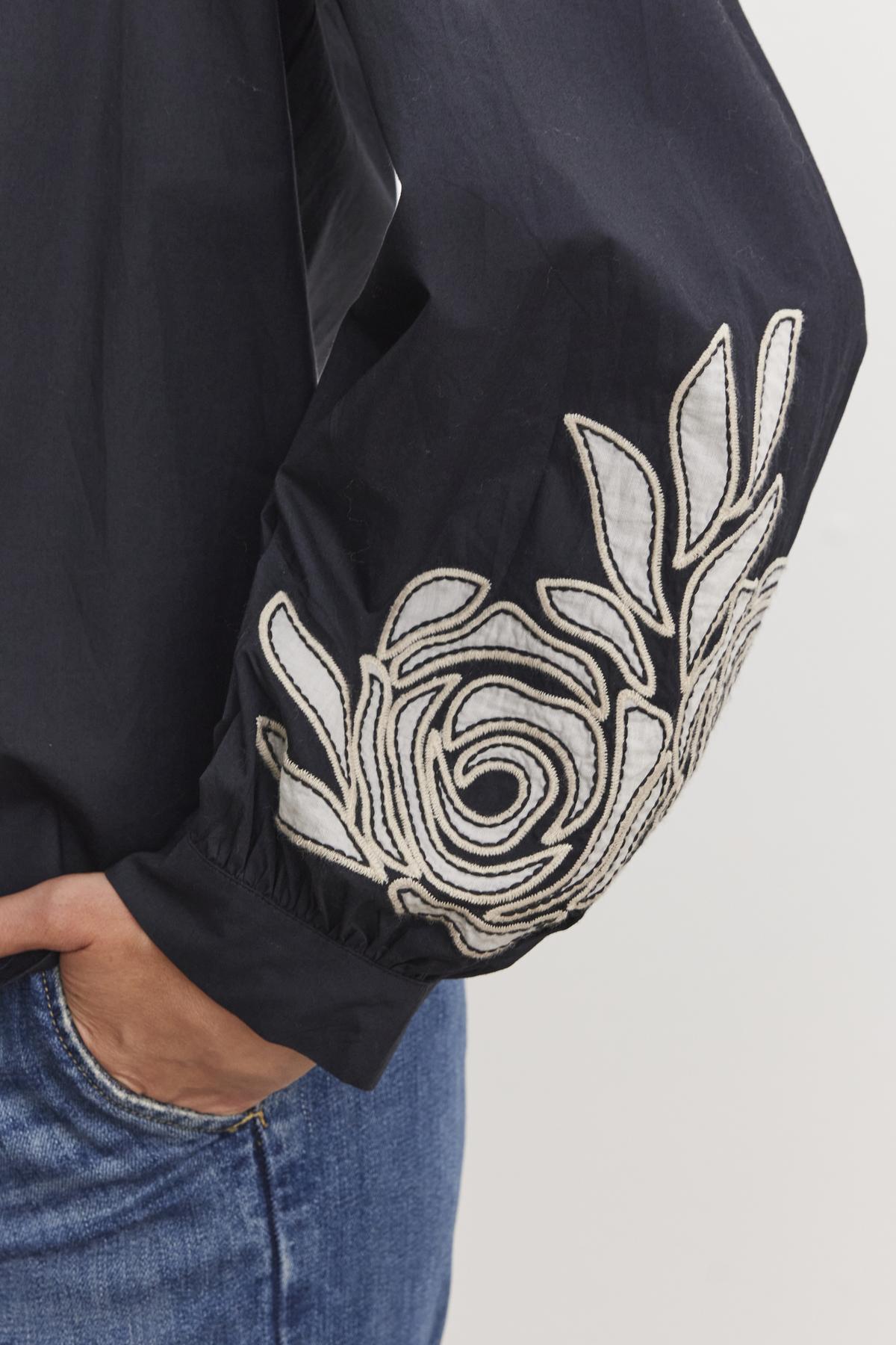 A close-up shows a person wearing the MARTINE TOP by Velvet by Graham & Spencer, a black cotton poplin top with two-tone floral embroidery on the sleeve, paired with blue jeans.