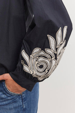 A close-up shows a person wearing the MARTINE TOP by Velvet by Graham & Spencer, a black cotton poplin top with two-tone floral embroidery on the sleeve, paired with blue jeans.