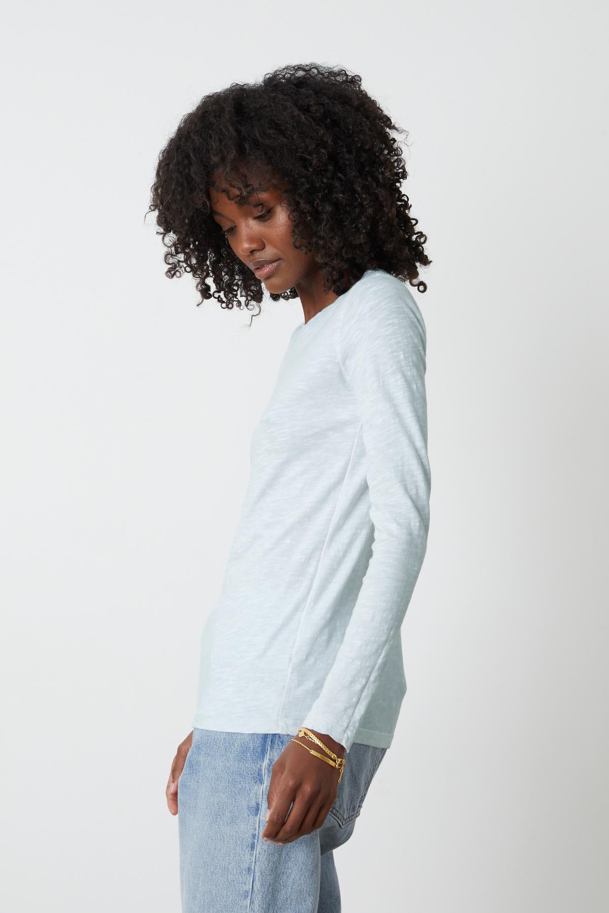 A person with curly hair is wearing a light blue long-sleeve LIZZIE TEE by Velvet by Graham & Spencer and denim jeans. They are standing sideways, looking down.-35982858780865