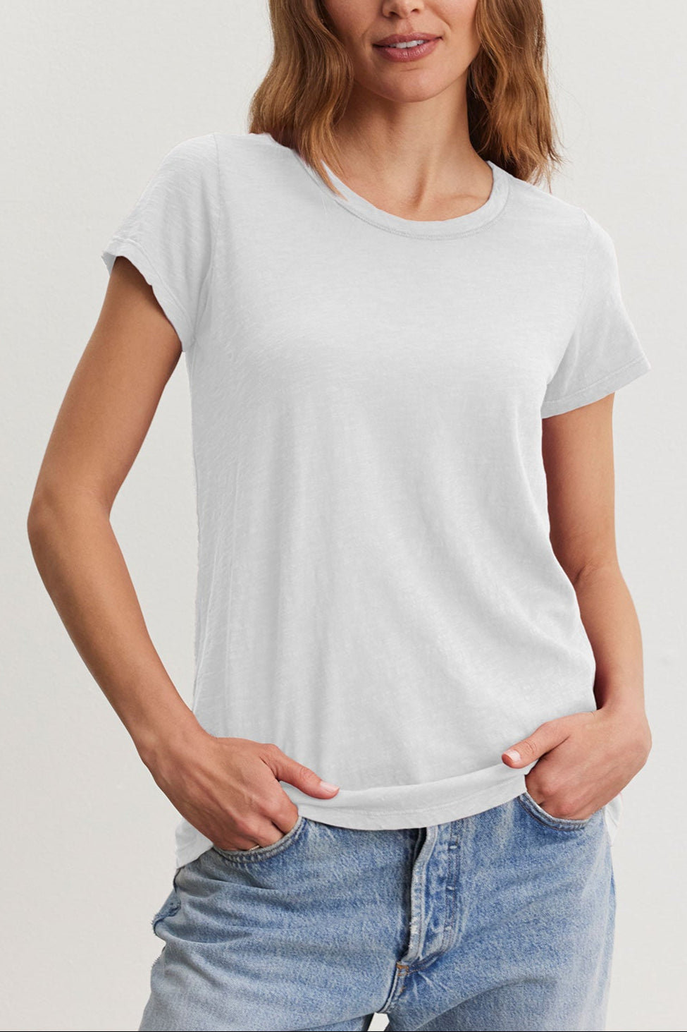 Wearing the TILLY TEE by Velvet by Graham & Spencer, a woman with wavy hair showcases effortless style in a California classic. She pairs it with blue jeans, standing with hands in pockets against a neutral background.-38942787403969
