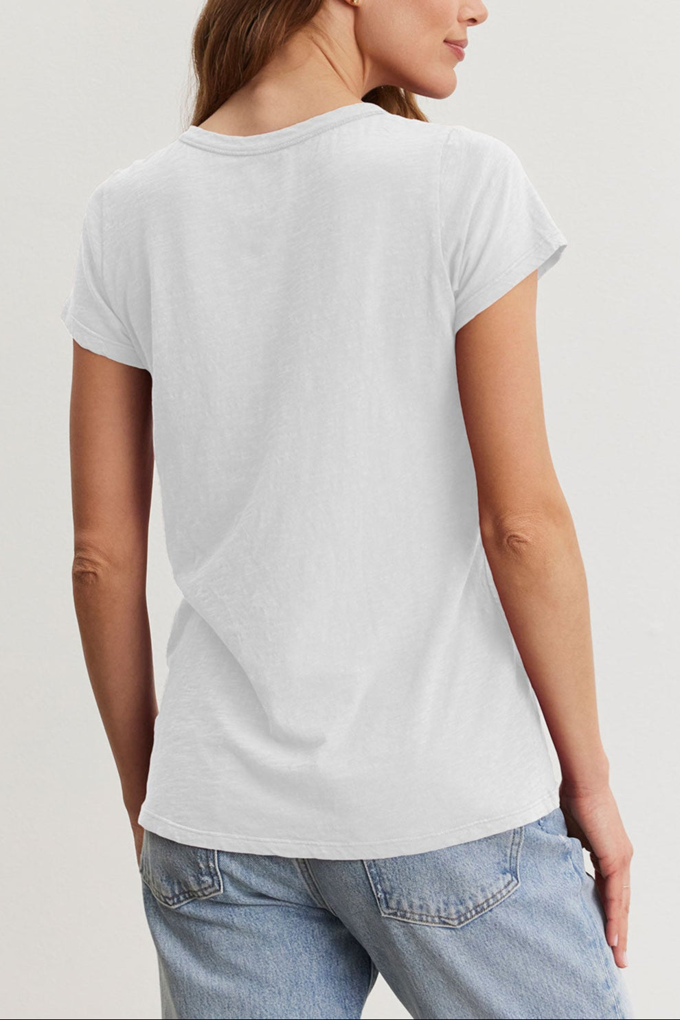 A person with long hair stands facing away in a California classic style, wearing a crisp, TILLY TEE by Velvet by Graham & Spencer and jeans against a plain background.-38942787436737