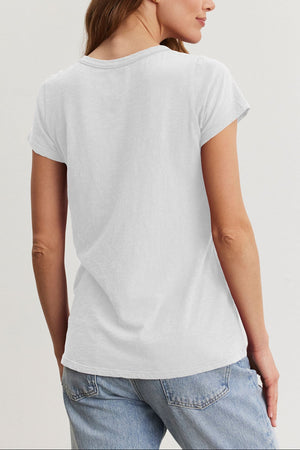 A person with long hair stands facing away in a California classic style, wearing a crisp, TILLY TEE by Velvet by Graham & Spencer and jeans against a plain background.