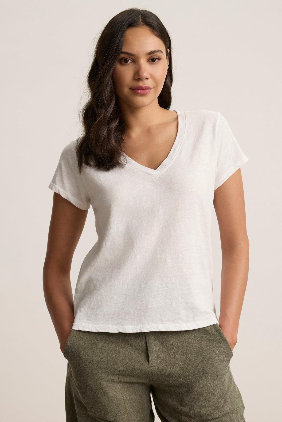 Wearing the relaxed-fit ELI TEE, a white cotton slub V-neck by Velvet by Graham & Spencer, and green pants, a person stands with hands in pockets against a neutral background.