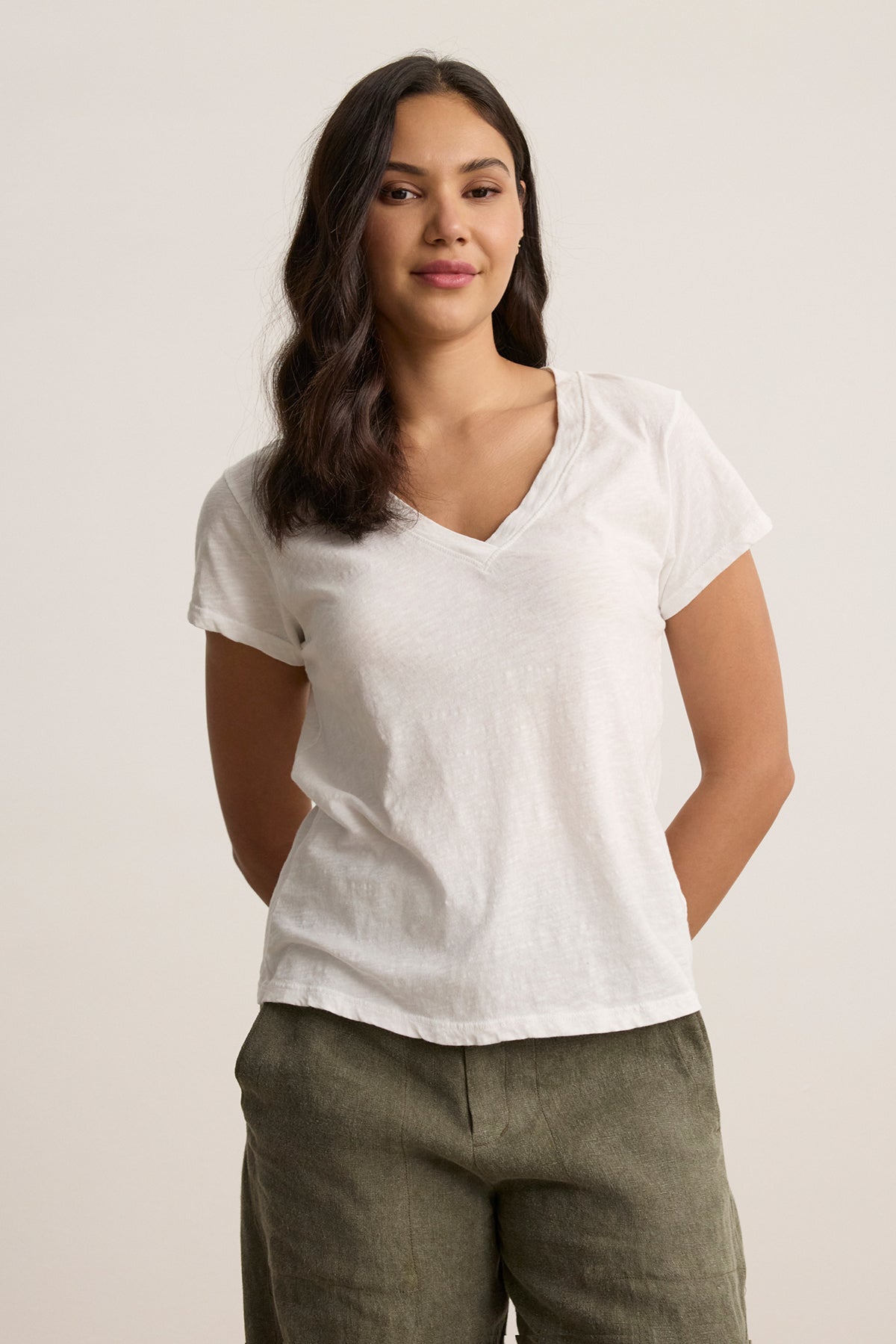 A person wearing the ELI TEE, a textured white V-neck from Velvet by Graham & Spencer, paired with green pants, stands against a plain background with hands behind their back.-38745911001281