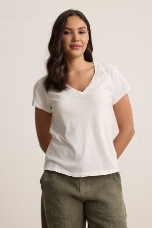A person wearing the ELI TEE, a textured white V-neck from Velvet by Graham & Spencer, paired with green pants, stands against a plain background with hands behind their back.
