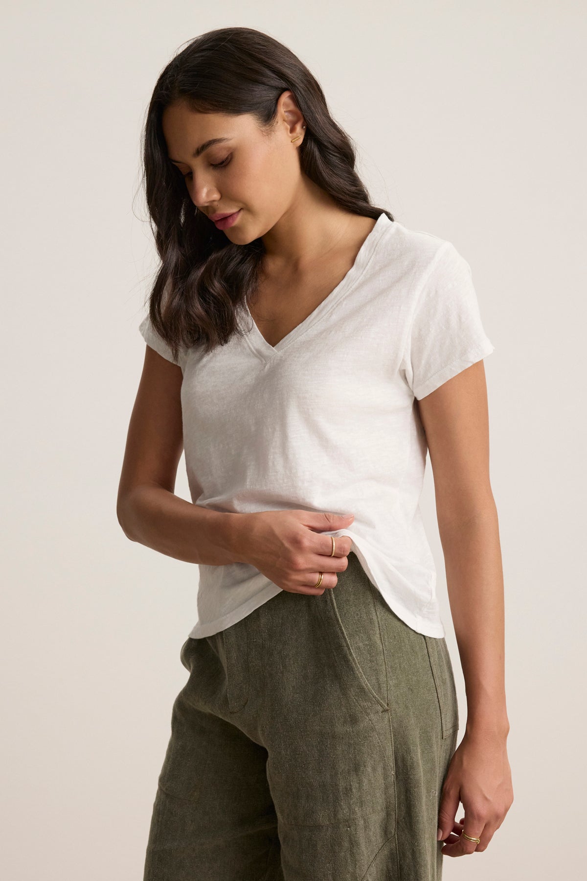 A woman in a white ELI TEE from Velvet by Graham & Spencer, featuring a relaxed fit cotton slub V-neck, paired with green pants, stands against a plain background looking downward.