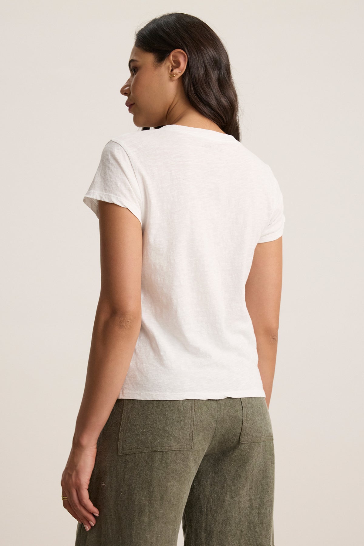 Someone wearing the ELI TEE, a white cotton slub V-neck from Velvet by Graham & Spencer, paired with green pants, stands looking aside. The relaxed fit blends seamlessly with the plain background.-38745910411457
