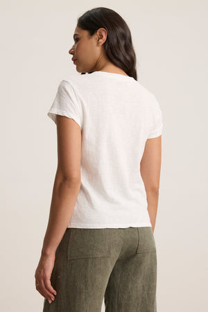 Someone wearing the ELI TEE, a white cotton slub V-neck from Velvet by Graham & Spencer, paired with green pants, stands looking aside. The relaxed fit blends seamlessly with the plain background.