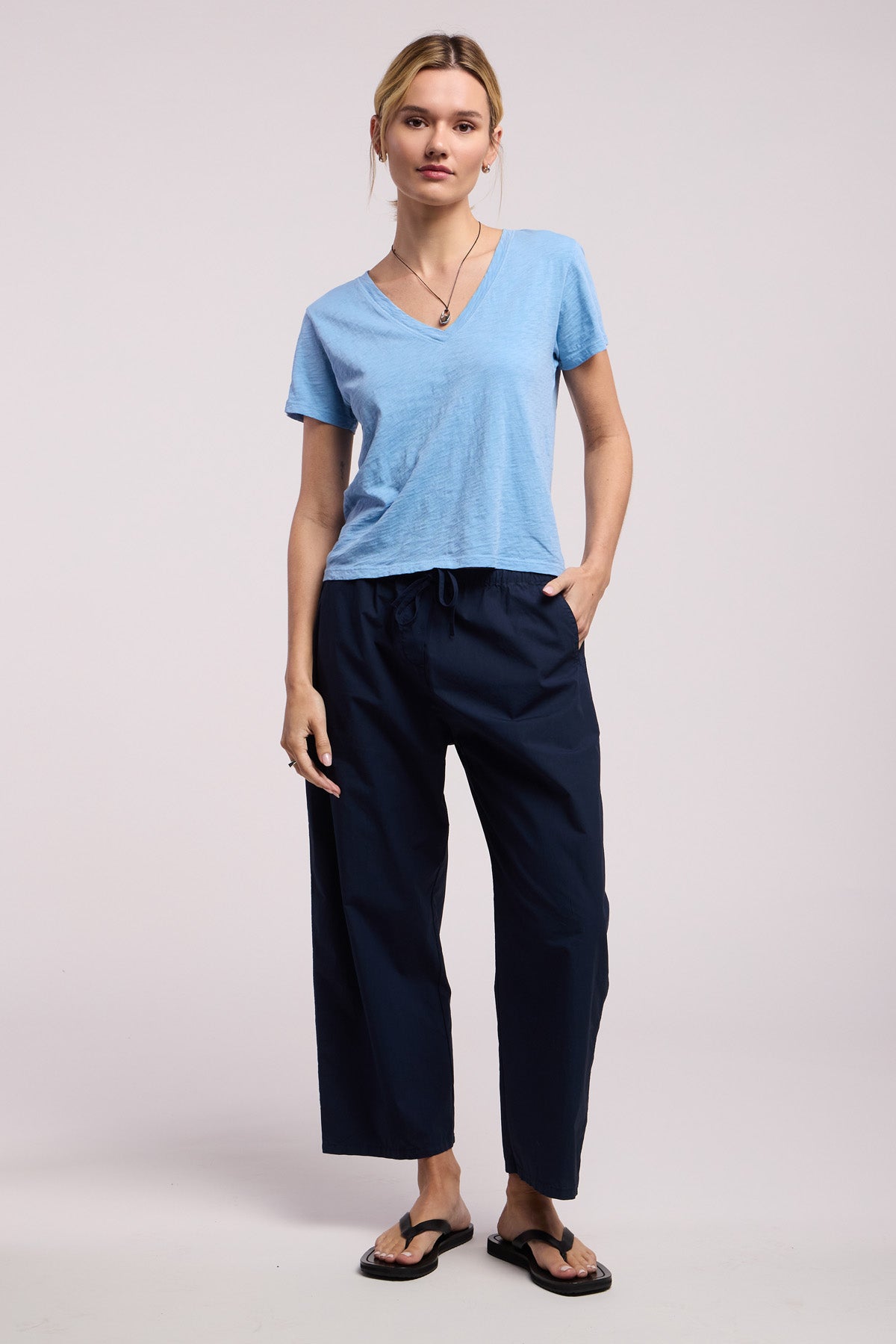 Wearing the ELISHA TEE by Velvet by Graham & Spencer—a light blue cotton slub V-neck—paired with dark blue loose pants, black flip flops, and a necklace, one hand in pocket against a plain background; a true wardrobe staple.-41645010845889