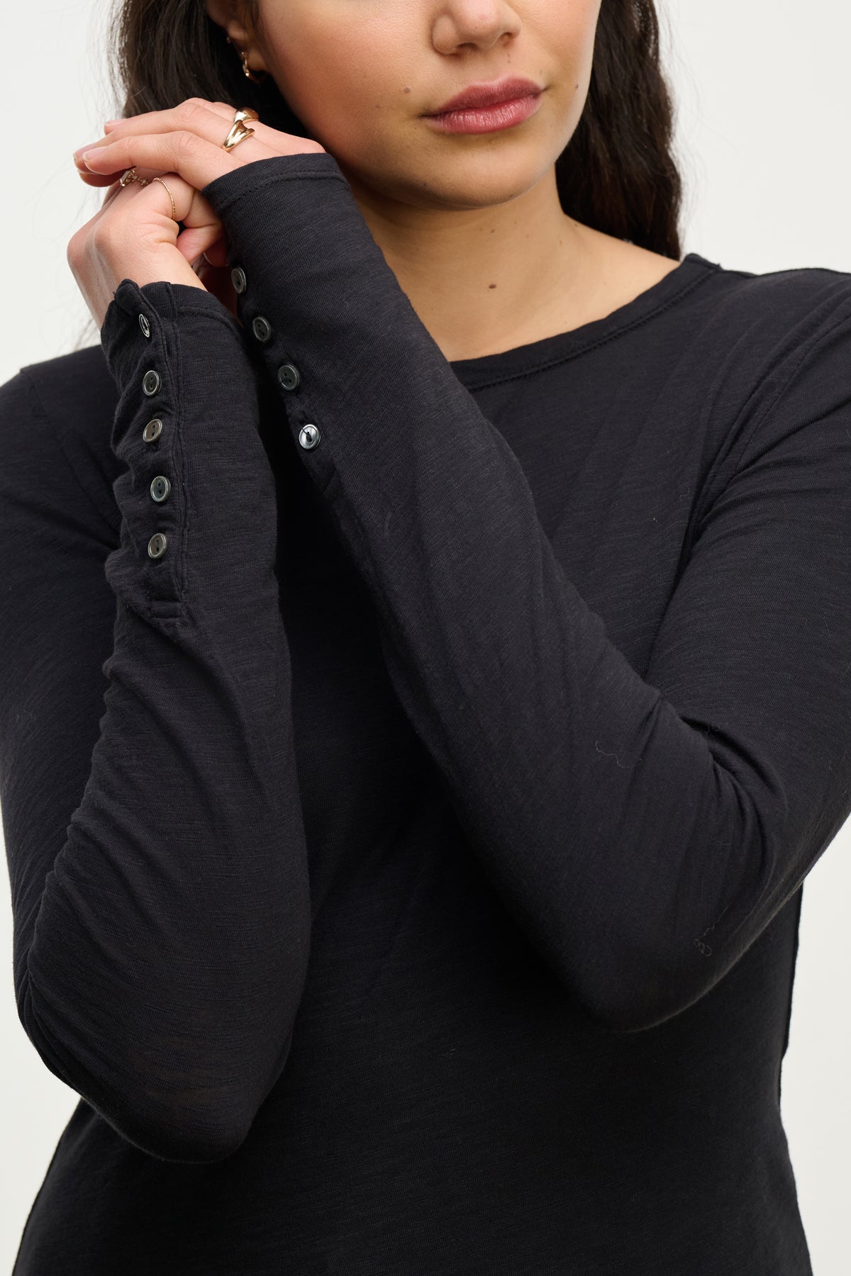 A woman wearing the KELSEY TEE by Velvet by Graham & Spencer, black with button sleeves, has her hands clasped near her face and lips partially visible.