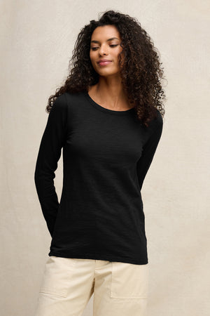 A person with curly hair wears the LIZZIE TEE by Velvet by Graham & Spencer—this long-sleeve, crew-neck cotton slub top is paired with light-colored pants against a neutral background.