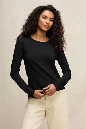 A woman with curly hair wears the LIZZIE TEE by Velvet by Graham & Spencer, a black cotton slub crew neck top, paired with light pants and standing against a plain beige background.