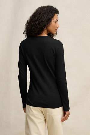 Someone with curly hair, seen from behind against a neutral background, wears the LIZZIE TEE by Velvet by Graham & Spencer—a long-sleeve black cotton slub crewneck—paired with beige pants.