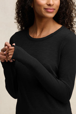 A woman with curly hair wears the Velvet by Graham & Spencer LIZZIE TEE, a long sleeve black cotton slub crewneck, standing with her hands clasped and facing slightly to the side.