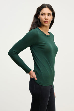 A woman stands in profile against a light background, wearing the LIZZIE TEE by Velvet by Graham & Spencer—a long sleeve, dark green cotton slub crew neck tee—paired with dark jeans.
