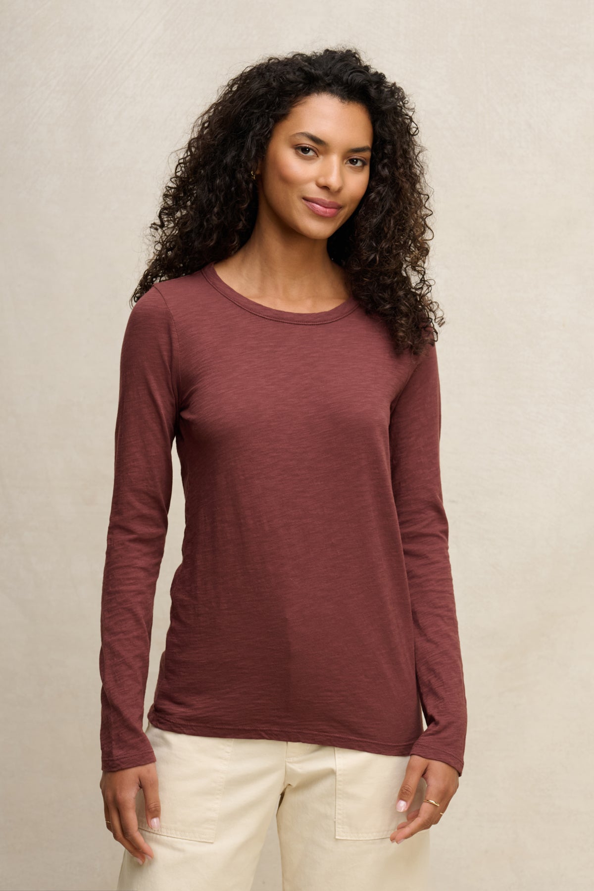 A woman with curly hair wears the Velvet by Graham & Spencer LIZZIE TEE—long sleeves, crew neckline, maroon color—with light pants, standing against a plain light background.