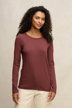 A woman with curly hair wears the Velvet by Graham & Spencer LIZZIE TEE—long sleeves, crew neckline, maroon color—with light pants, standing against a plain light background.