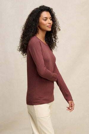 A woman with curly hair stands sideways against a beige background, wearing the Velvet by Graham & Spencer LIZZIE TEE—a maroon, long sleeve crewneck—paired with light-colored pants.