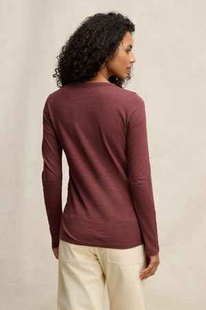 A person with curly hair wears the Velvet by Graham & Spencer LIZZIE TEE, a maroon long sleeve crew neck, paired with cream pants, and stands facing away from the camera against a neutral background.