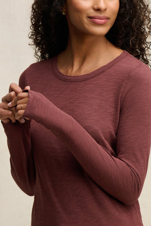 A woman with curly hair models the LIZZIE TEE by Velvet by Graham & Spencer, a mauve, long sleeve crew neck top, standing with her hands folded and facing partially away from the camera.