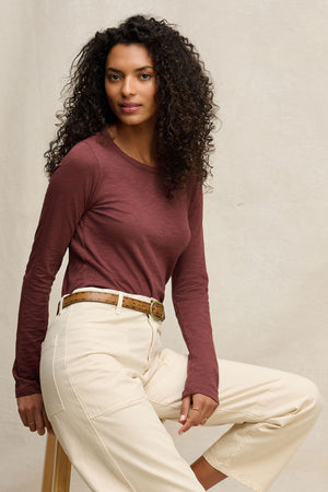 A woman with curly hair wears the Velvet by Graham & Spencer LIZZIE TEE in maroon, featuring a cotton slub fabric and crew neck, paired with beige high-waisted pants and a brown belt, seated on a wooden stool against a neutral background.
