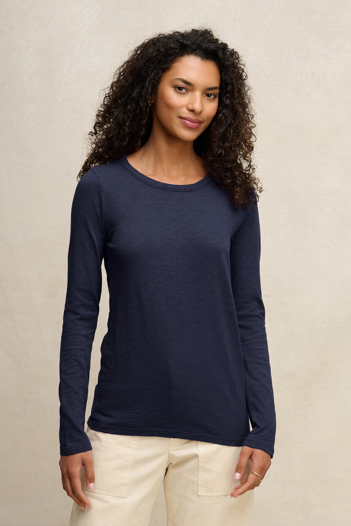 A woman with curly hair wears the Velvet by Graham & Spencer LIZZIE TEE, a long-sleeve navy blue crew neck shirt, paired with light-colored pants against a plain, light background.