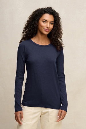 A woman with curly hair wears the Velvet by Graham & Spencer LIZZIE TEE, a long-sleeve navy blue crew neck shirt, paired with light-colored pants against a plain, light background.