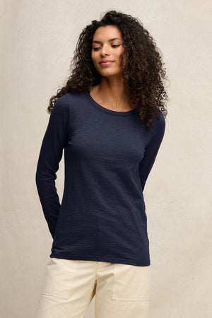 A woman with curly hair models the LIZZIE TEE by Velvet by Graham & Spencer, a navy cotton slub crewneck with long sleeves, paired with light pants. She stands against a neutral backdrop, gazing slightly to the side, hands behind her back.