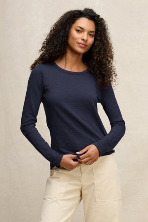 A woman with curly hair wears the Velvet by Graham & Spencer LIZZIE TEE, a long sleeve navy blue cotton slub top with a crew neckline, paired with light beige pants against a plain light background.