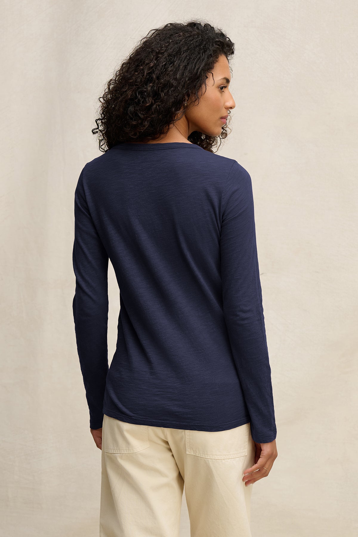 A person with curly hair is seen from the back wearing the Velvet by Graham & Spencer LIZZIE TEE in navy blue with long sleeves and cream-colored pants, set against a plain light background.