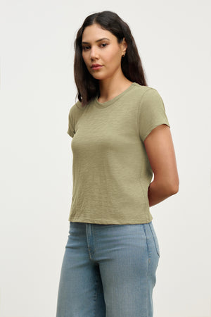 A woman with long dark hair models the MARIE TEE by Velvet by Graham & Spencer—a light green, relaxed boxy casual top—paired with blue jeans, standing against a plain light background with her hands behind her back.