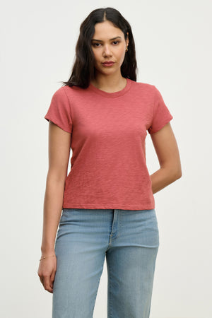 A woman with long dark hair wears the MARIE TEE by Velvet by Graham & Spencer—a relaxed, boxy-fit pink cotton slub t-shirt—paired with light blue jeans, standing against a plain white background. Perfect for any casual occasion.