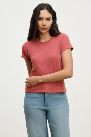 A woman with long dark hair wears the MARIE TEE from Velvet by Graham & Spencer—a short-sleeved, relaxed boxy fit red cotton slub t-shirt—with light blue jeans, standing against a plain light background.