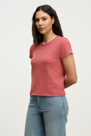 A woman with long dark hair models the MARIE TEE by Velvet by Graham & Spencer—a relaxed, boxy-fit pink short-sleeve cotton slub shirt—paired with light blue jeans against a plain background, ideal for casual wear.
