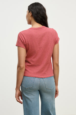 A woman faces away from the camera, wearing the MARIE TEE by Velvet by Graham & Spencer—a short-sleeve red cotton slub top—paired with relaxed-fit light blue jeans, ideal for casual occasions against a plain background.