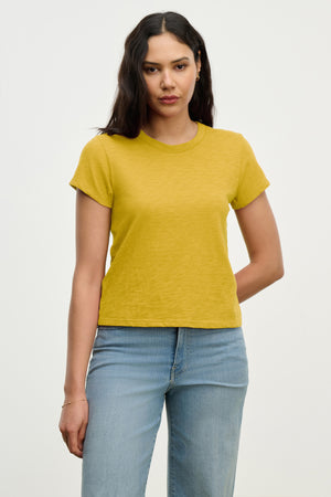 A woman with long dark hair wears the MARIE TEE by Velvet by Graham & Spencer, a yellow casual top with a relaxed boxy fit, styled with light blue jeans against a plain light background.