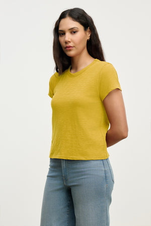 A woman with long dark hair wears the MARIE TEE by Velvet by Graham & Spencer in yellow and light blue jeans, standing with hands behind her back against a plain light background.