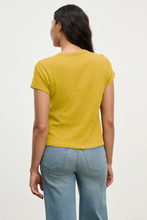A woman faces away wearing the MARIE TEE by Velvet by Graham & Spencer, a relaxed fit yellow casual tee paired with light blue jeans against a plain background.