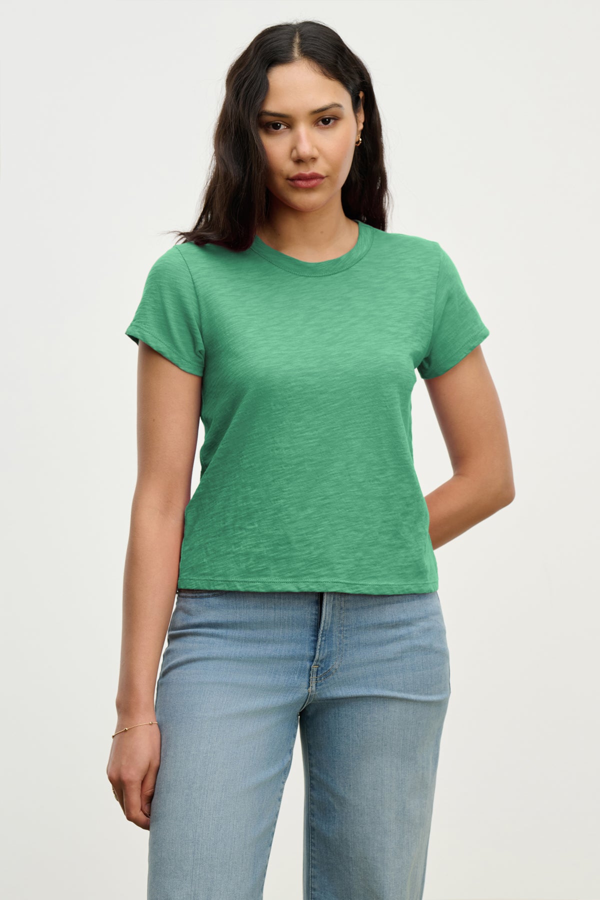 A woman with long dark hair wears the MARIE TEE by Velvet by Graham & Spencer in green cotton slub, paired with light blue jeans against a plain backdrop—ideal for any casual occasion.-39319728685249