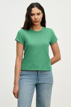 A woman with long dark hair wears the MARIE TEE by Velvet by Graham & Spencer in green cotton slub, paired with light blue jeans against a plain backdrop—ideal for any casual occasion.