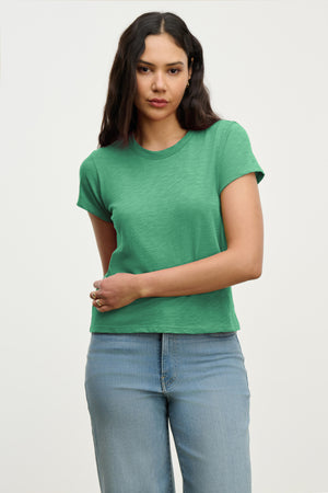 Woman with long dark hair wears the MARIE TEE by Velvet by Graham & Spencer—a green cotton slub short-sleeve t-shirt in a relaxed, boxy fit—paired with light blue jeans, standing against a plain light background.