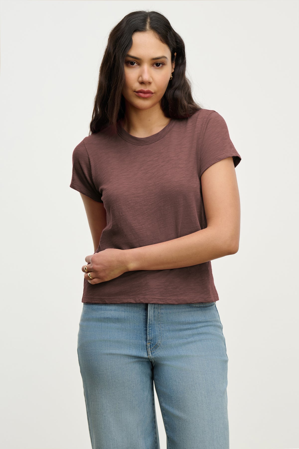 A woman with long dark hair wears the MARIE TEE by Velvet by Graham & Spencer—a short-sleeve brown, relaxed boxy fit t-shirt—with light blue jeans, standing against a plain light background.-39319729537217
