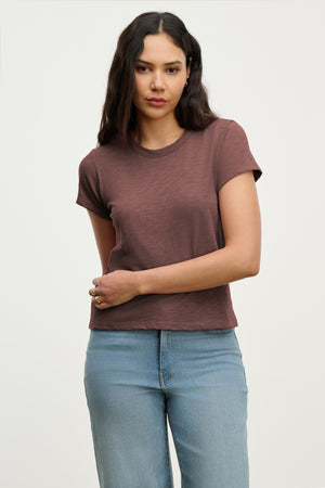 A woman with long dark hair wears the MARIE TEE by Velvet by Graham & Spencer—a short-sleeve brown, relaxed boxy fit t-shirt—with light blue jeans, standing against a plain light background.